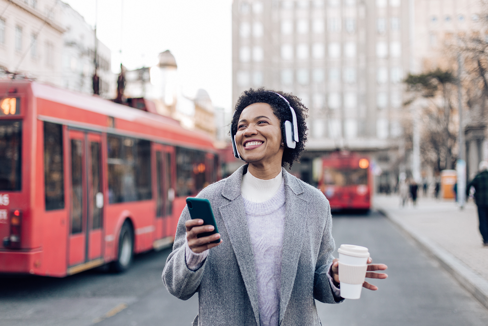 smiling woman walking through town, listening to music through headphones and holding coffee