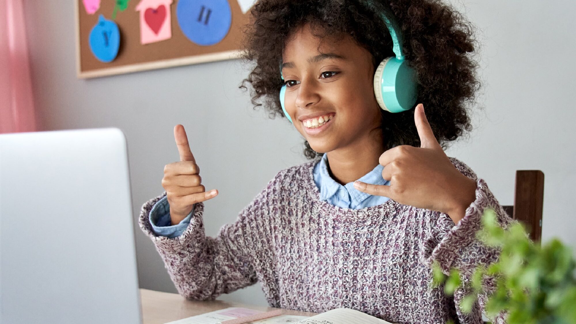 child sitting at desk with headphones on smiling and making gesture at laptop