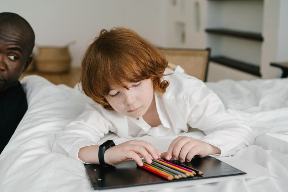Ginger young boy lying on bed on his stomach arranging coloured pencils while man looks on at him