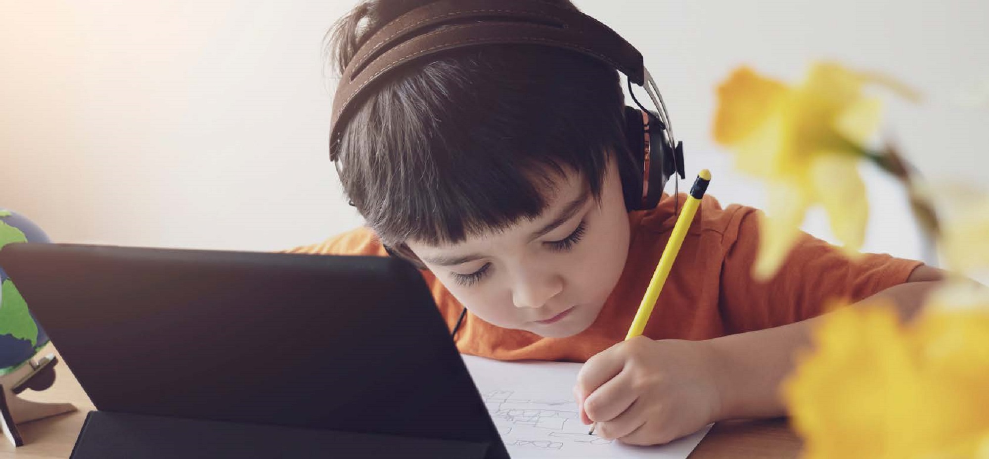 young boy in front of a tablet with headphones on writing with a pencil