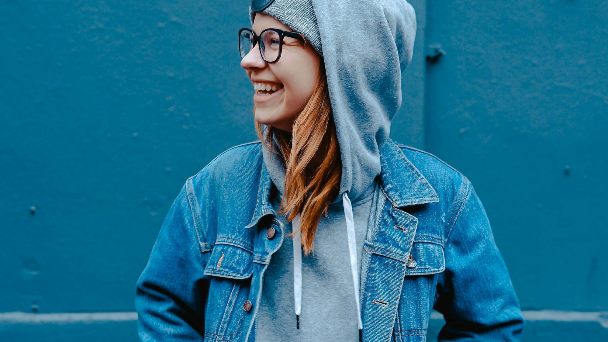 Smiling young woman in glasses and denim jacket layered over grey hoodie against blue urban wall