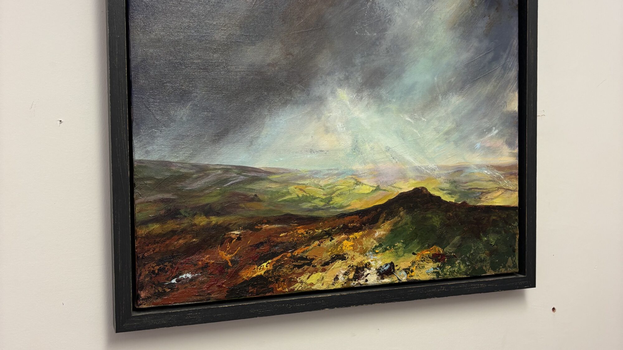 Framed oil landscape painting on a white wall showing a stormy sky above vibrant green hills and textured foreground