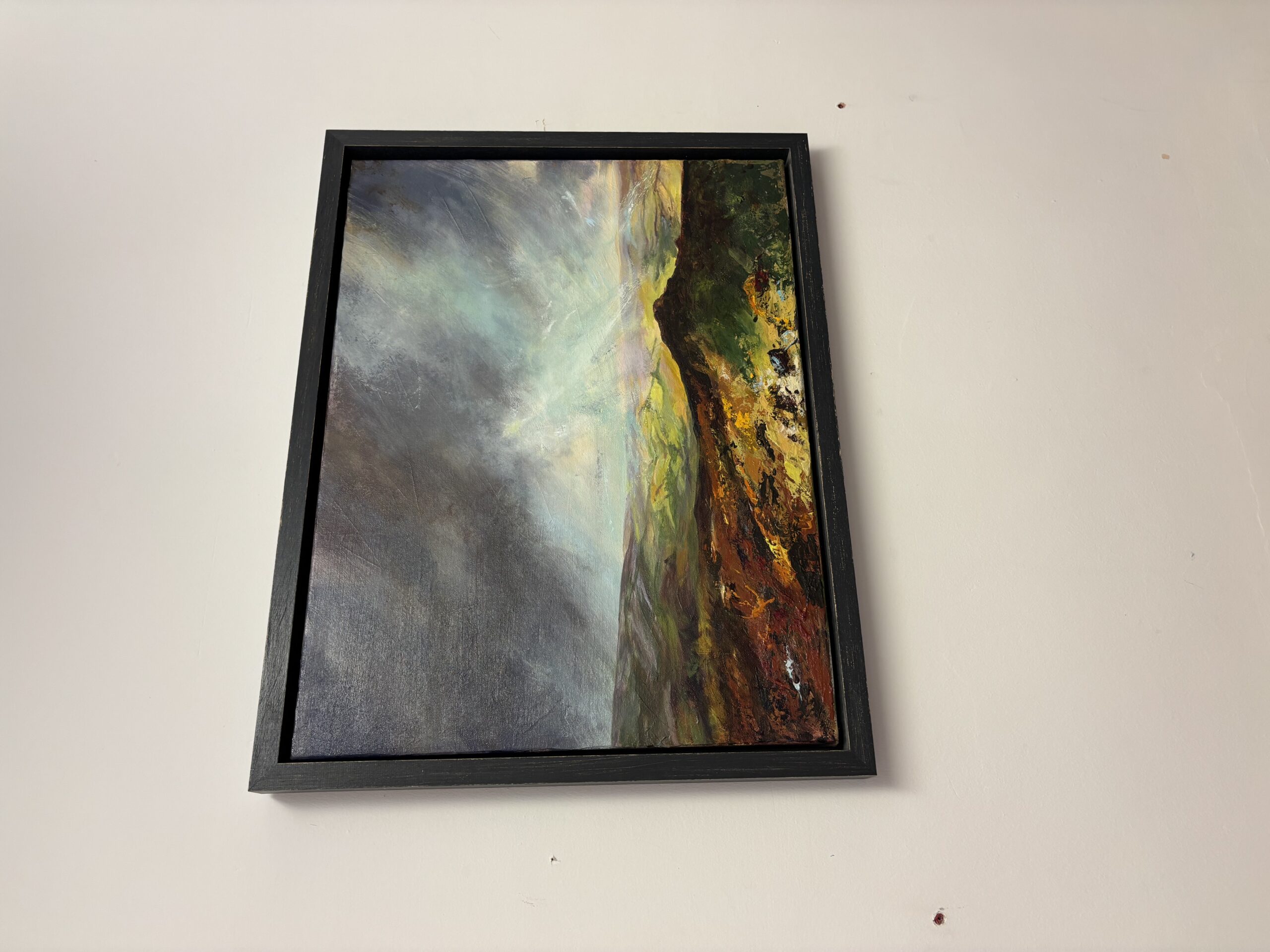 Framed oil landscape painting on a white wall showing a stormy sky above vibrant green hills and textured foreground