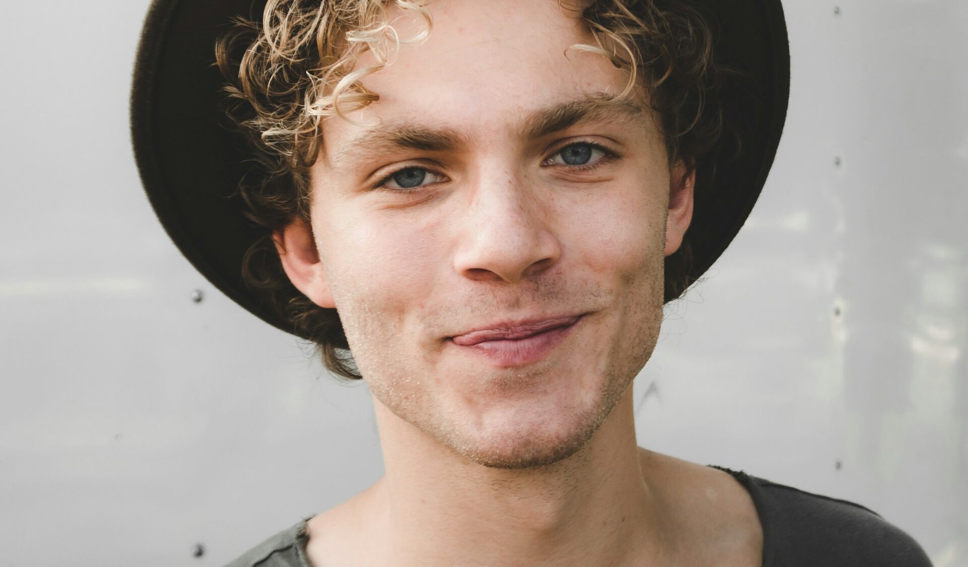 Smiling young man close-up portrait with curly hair, black hat and casual dark shirt, lifestyle headshot