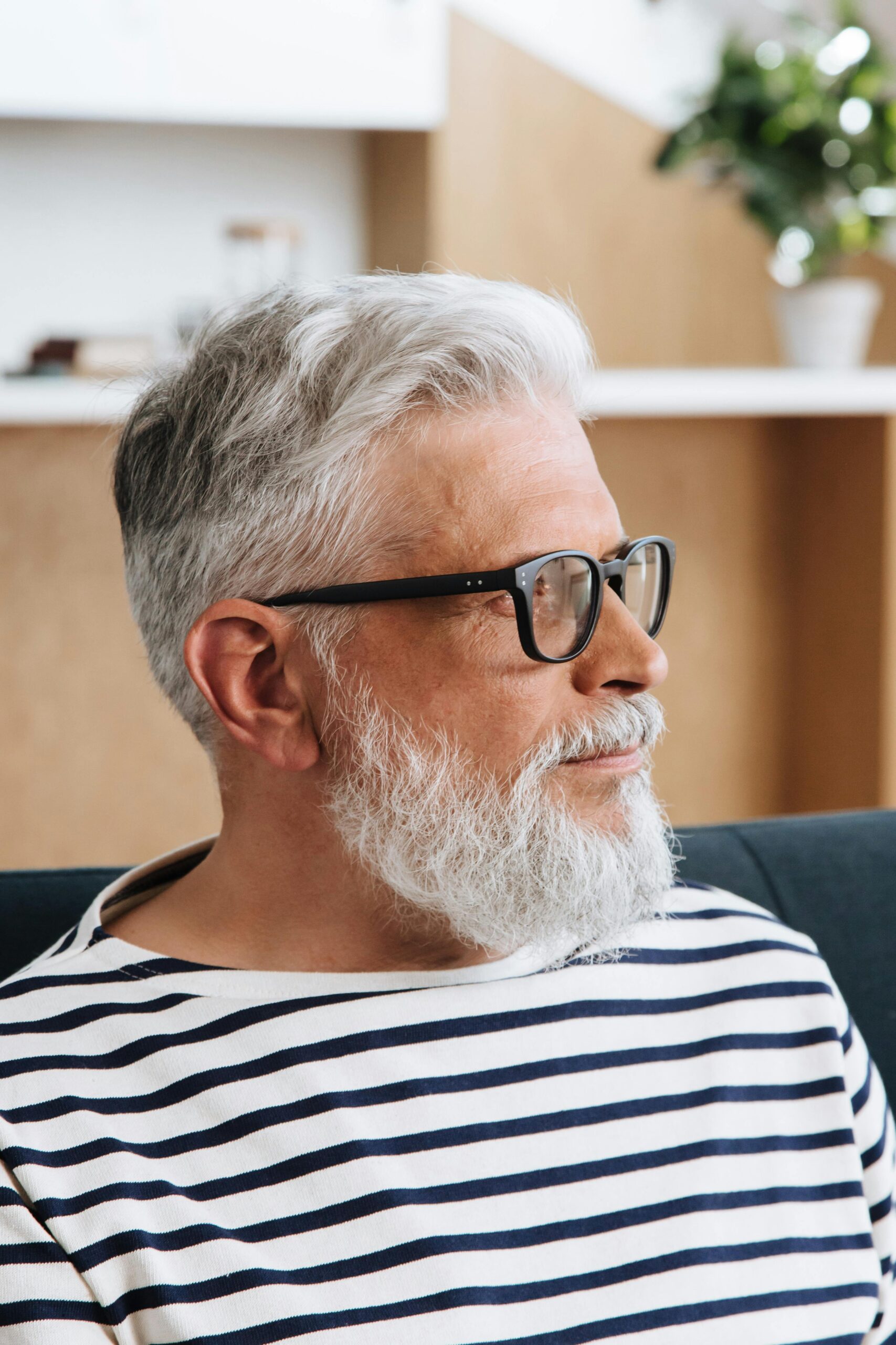 Senior man with gray hair and beard wearing black glasses and striped shirt, side-profile portrait in a modern home.