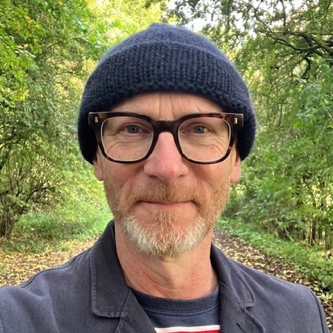 Smiling man wearing glasses and navy beanie walking on a wooded path surrounded by green trees and fallen leaves