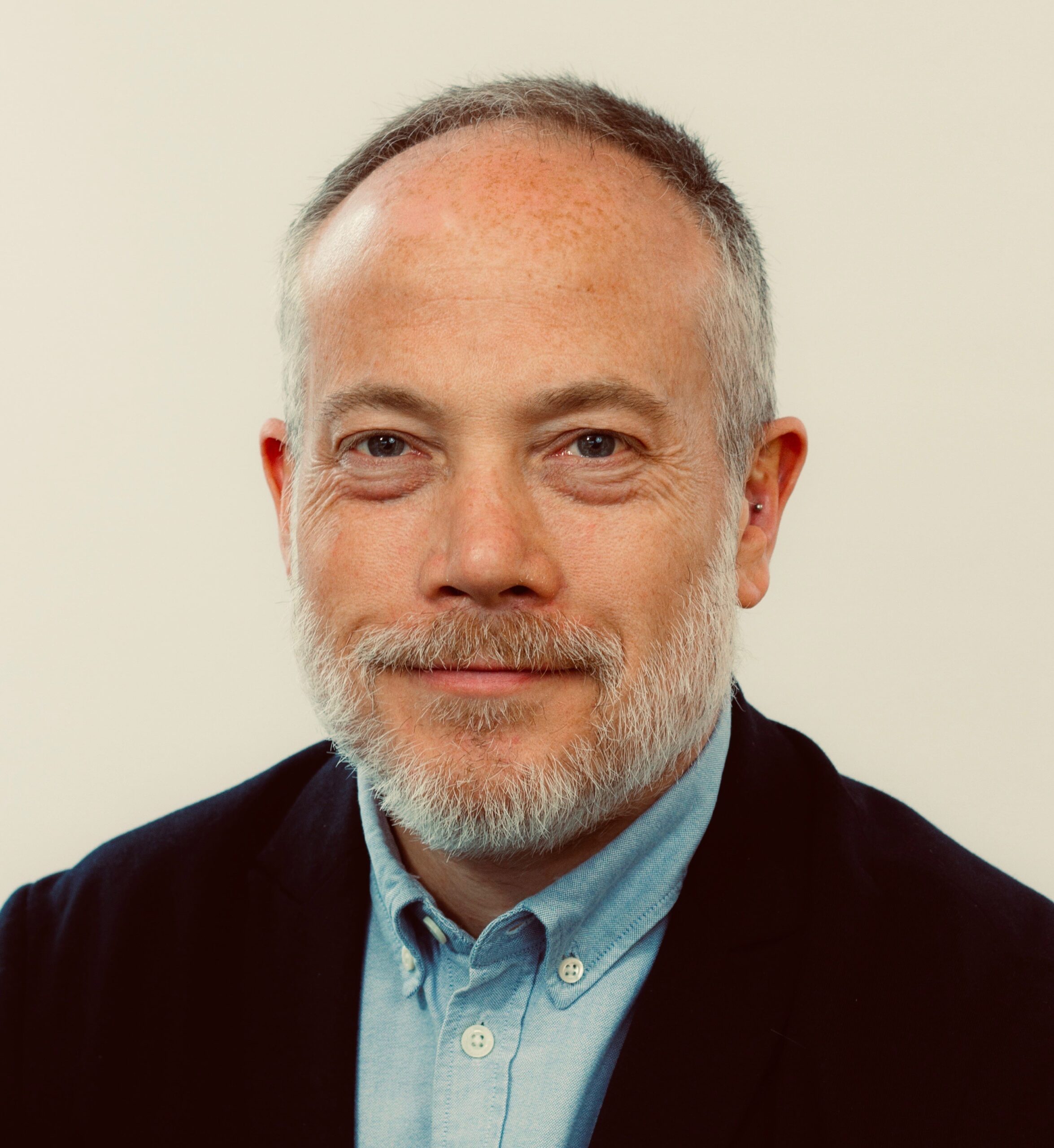 Close-up headshot of smiling middle-aged man with gray beard, receding hairline, wearing blue shirt and dark blazer