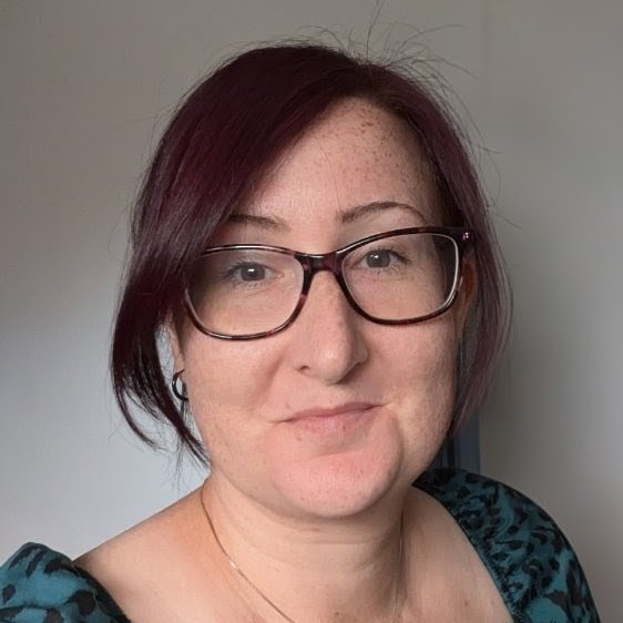 Portrait of smiling woman with burgundy hair and glasses in teal patterned top, neutral background